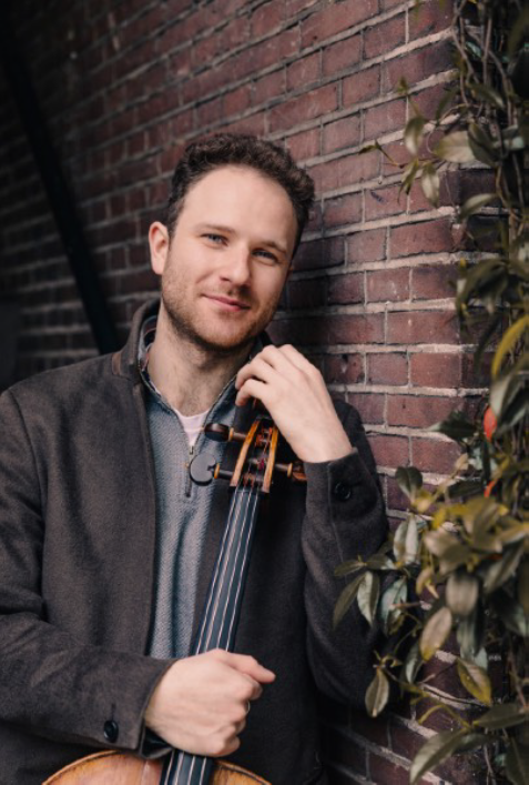 Image of white man holding a cello standing next to a brick wall